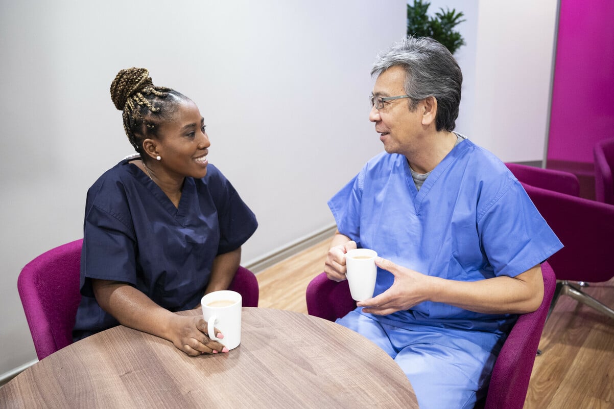 Two healthcare workers sitting in a break room having a coffee. They look calm and happy.
